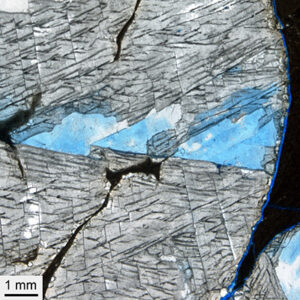 This photograph shows 245 million-year-old rock salt crystals (halite) at comparatively low magnification, embedded in dark mudstone. The lines and chevrons mark the crystal axes. The width of the sample is 10 mm.