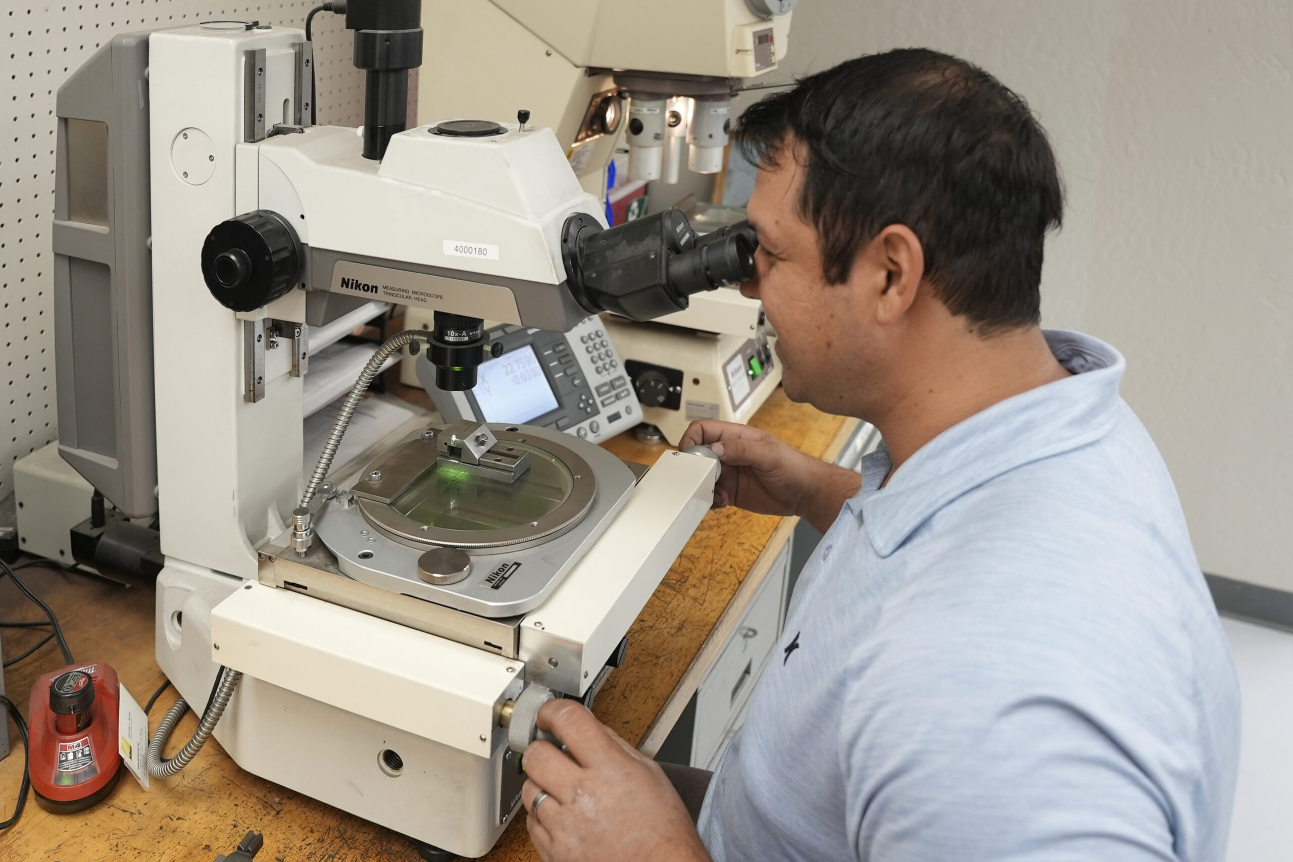 Teknisi sedang melakukan inspeksi optik pada komponen hasil stamping menggunakan Measuring Microscope dari Nikon.